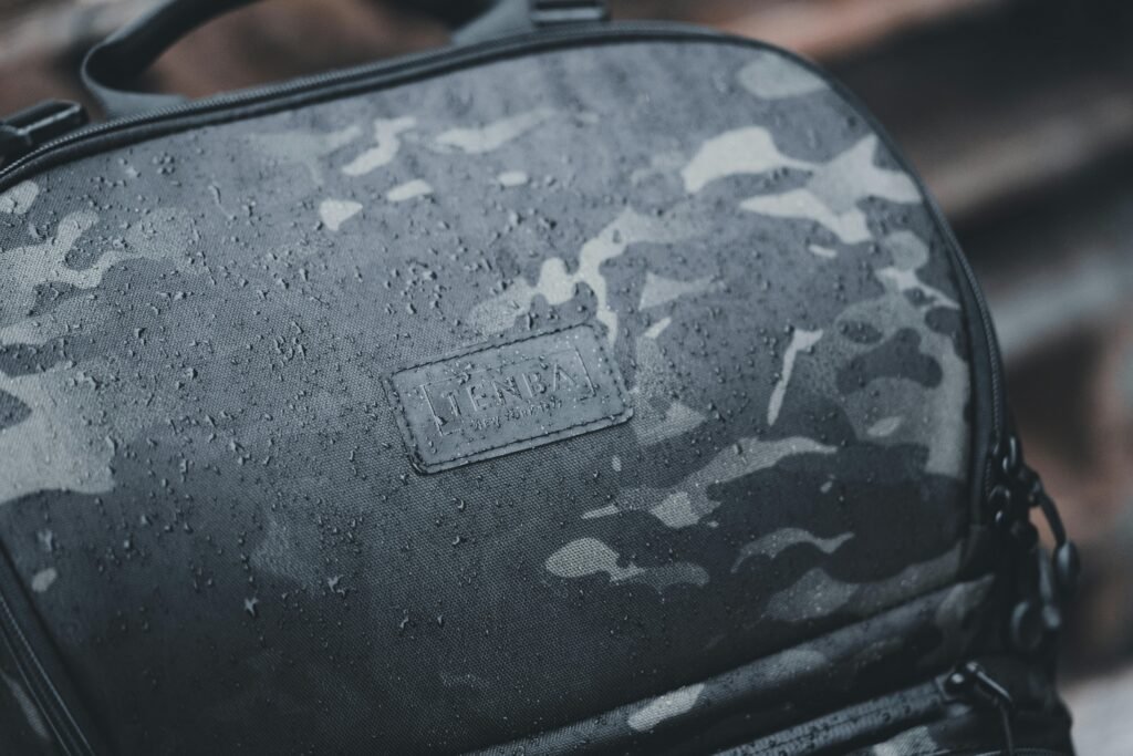 pexels-photo-16359260-16359260 Close-up of a waterproof backpack with a camouflage pattern and branded logo, wet from rain.